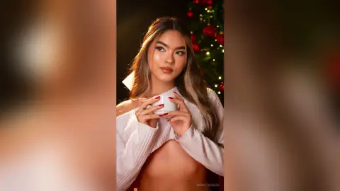 Young woman in a white off-the-shoulder sweater holding a coffee cup near a festive Christmas tree.