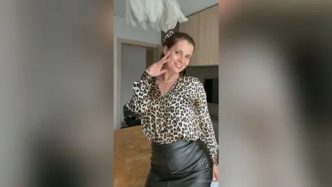 Smiling woman in a leopard print blouse and black leather skirt posing in a modern minimalist living room.
