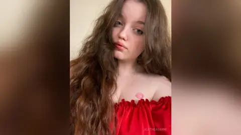 Young woman with long wavy hair wearing a red off-the-shoulder velvet top in a soft indoor setting.