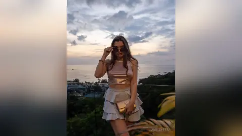 Stylish woman in a champagne satin outfit and sunglasses posing against a sunset ocean view.