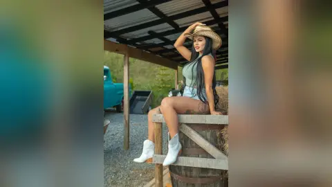 ErikaHoffman sits on a wooden barrel wearing a light green tank top, denim shorts, and a straw hat, posing with one hand on her head.