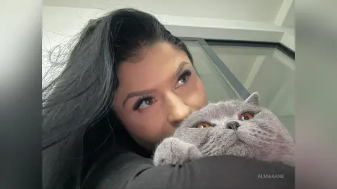 AlmaKane — Young woman with long dark hair cuddling a grey British Shorthair cat in a cozy indoor setting.