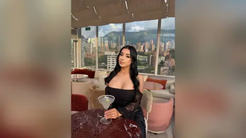 Elegant woman in a black lace dress enjoying a cocktail at a luxury rooftop lounge with a city skyline view.