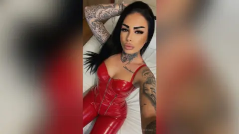 Tattooed woman in a red latex bodysuit lounging on a white bed with a confident mood.