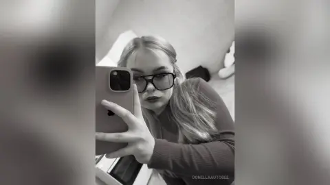 Young woman with blonde hair and glasses taking a moody selfie in a long-sleeved top.