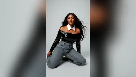 Elegant Black woman in a black off-the-shoulder top and grey jeans posing against a grey studio background.