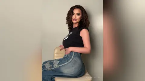 Curvy woman in a black graphic t-shirt and distressed denim jeans sitting on a chair against a neutral wall.