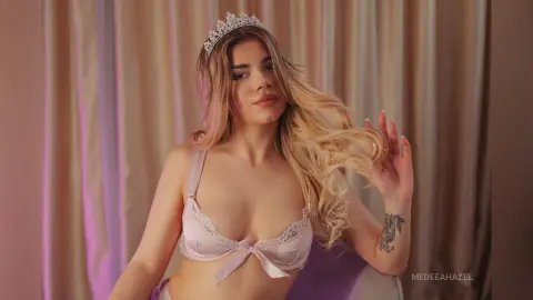 Blonde woman in white lace lingerie and a silver tiara posing glamorously against soft draped curtains.