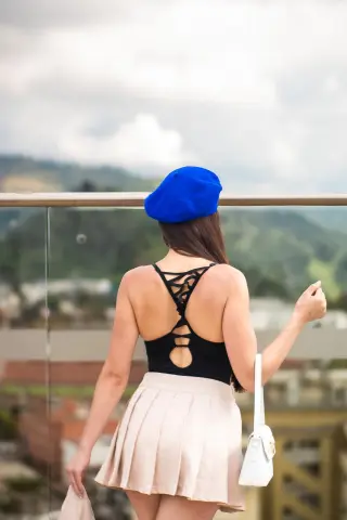 Woman in a blue beret and beige pleated skirt standing on a balcony overlooking a scenic mountain landscape.