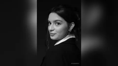 Portrait of a young woman with dark hair wearing a black blazer against a dark moody background.