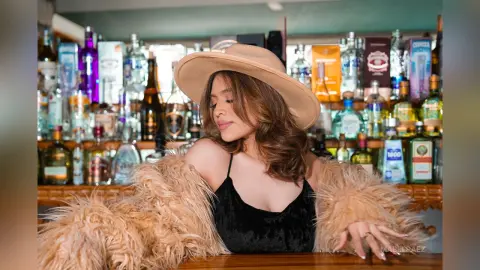 Stylish woman in a tan wide-brim hat and fuzzy coat posing in a moody, dimly lit bar setting.
