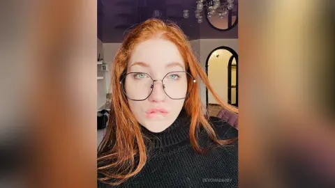 Redheaded woman with glasses in a black turtleneck sweater making a pouty face in a modern living room.
