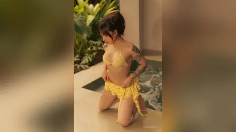 Young woman in a yellow bikini and matching sarong kneeling by a tropical poolside garden.