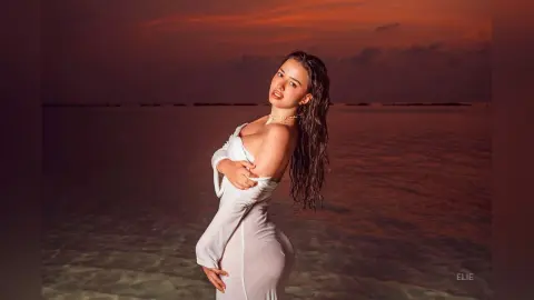 Elie poses in a white dress against a sunset ocean background, looking back over her shoulder with wet hair.