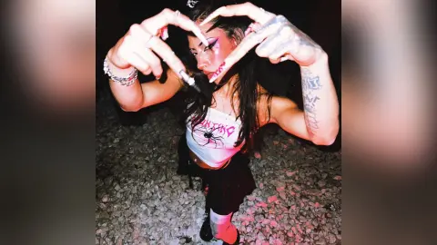 LiaValmont poses with her hands framing her face, wearing a white tank top and black skirt against a dark, textured ground.