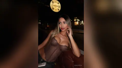 MiaGuerra poses in a sheer, lacy brown corset, showing off her cleavage and long blonde hair against a dark, outdoor night setting.