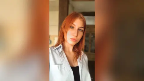 Redheaded woman in a white button-down shirt posing with a serene expression in a sunlit indoor setting.