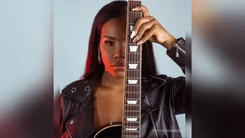 Stylish woman in a black leather jacket posing with an electric guitar in moody studio lighting.