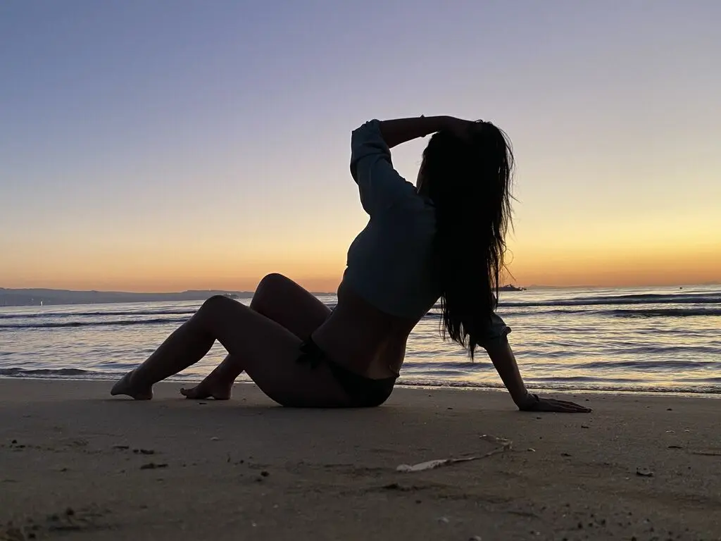 Silhouette of a woman in a crop top sitting on a sandy beach at sunset by the ocean waves.