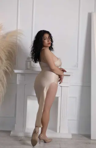 Elegant woman in a champagne satin slip dress with a high slit posing in a bright, minimalist white room.