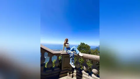 Woman in casual white top and blue jeans sitting on a stone balcony overlooking a scenic coastal city view.