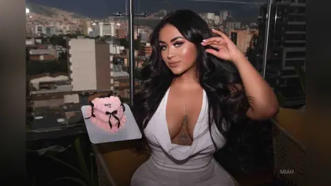 Glamorous woman in a white plunging dress holding a pink birthday cake on a city balcony at night.