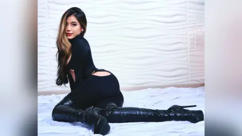 JessieLiu poses sitting on a white bed wearing a black long-sleeve bodysuit and black thigh-high leather boots.