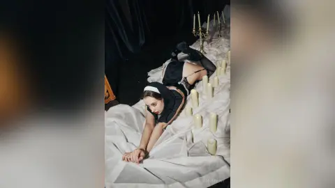 Woman in black lingerie lying on white sheets surrounded by glowing candles in a moody, dark setting.