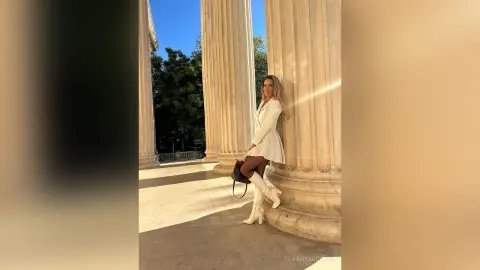 TiffanyAddams poses leaning against large stone columns, wearing a white mini dress and white boots with her blonde hair down.