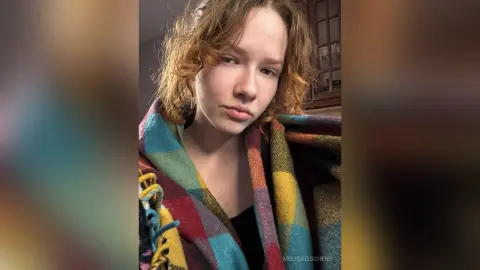 Young woman with curly hair wrapped in a colorful plaid blanket looking cozy in a dimly lit room.