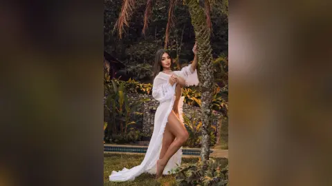 Elegant woman in a flowing white sheer robe posing by a palm tree in a lush tropical garden setting.