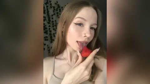 Young woman with freckles and winged eyeliner eating a strawberry in a cozy room with ivy decor.