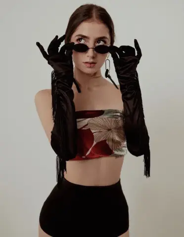 Elegant woman in floral tube top and long black satin gloves holding sunglasses against a neutral background.