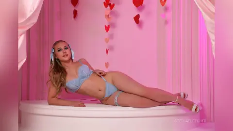 Blonde woman in light blue lace lingerie relaxing on a white platform in a dreamy pink studio setting.