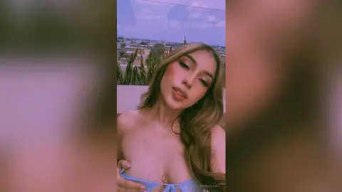 MeganLewit poses with long wavy hair, wearing a light blue bra that shows off her cleavage and bare chest against an outdoor balcony setting.