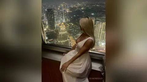Blonde woman in a white dress looking out at the glowing city skyline from a high-rise balcony at night.