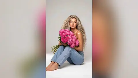 Blonde woman in denim jeans holding a large bouquet of pink peonies in a bright studio setting.