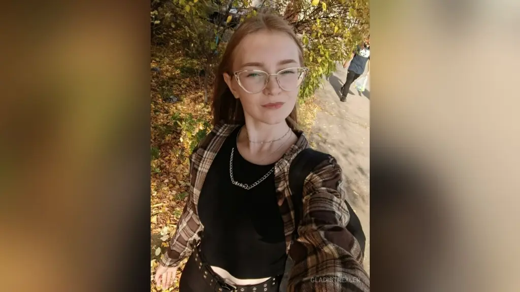 GladisTrexler β Young woman with glasses in a plaid shirt and black top smiling outdoors in a sunny autumn park setting.