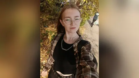 Young woman with glasses in a plaid shirt and black top smiling outdoors in a sunny autumn park setting.