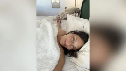 MichaelaBelle lies in bed with her dark hair spread on a white pillow, partially covered by a white textured blanket.