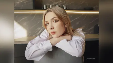 Blonde woman in a white shirt leaning on a chair in a modern kitchen with a soft, moody atmosphere.