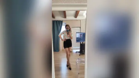 Young woman in a white blouse and black skirt posing for a mirror selfie in a bright, modern bedroom.