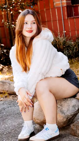 Smiling young woman in a cozy white faux fur jacket sitting outdoors in a sunny garden setting.