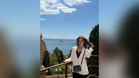 Stylish woman in a white blazer and sunglasses standing on a scenic balcony overlooking the ocean.