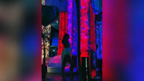 Aalyahh stands in a room with red brick walls and blue lighting, wearing a dark top and light-colored pants.