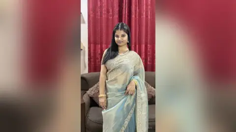Sejuti poses in a light blue and silver saree with gold jewelry, standing in front of red curtains.