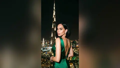 Elegant woman in a green satin evening gown posing against the glowing Dubai skyline at night.
