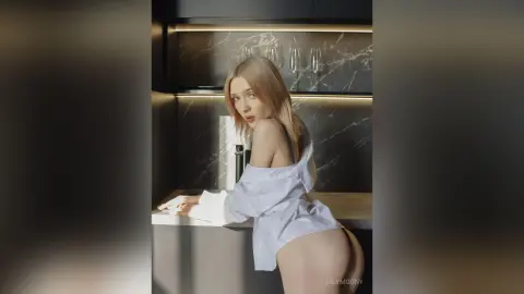 Blonde woman in an oversized white shirt posing in a modern kitchen with marble countertops and moody lighting.