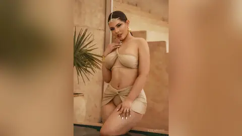 Elegant woman in a beige two-piece outfit and gold headpiece posing in a warm, sunlit Mediterranean setting.