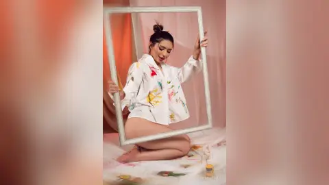 Young woman in a white paint-splattered shirt posing playfully with a large empty frame in a soft studio setting.